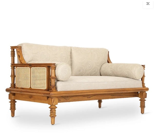A beautifully crafted wooden sofa set featuring intricate turned legs and natural cane webbing on the arms. This elegant sofa set includes comfortable, light-colored upholstered cushions with a subtle pattern and matching bolster pillows, all set against a white background.