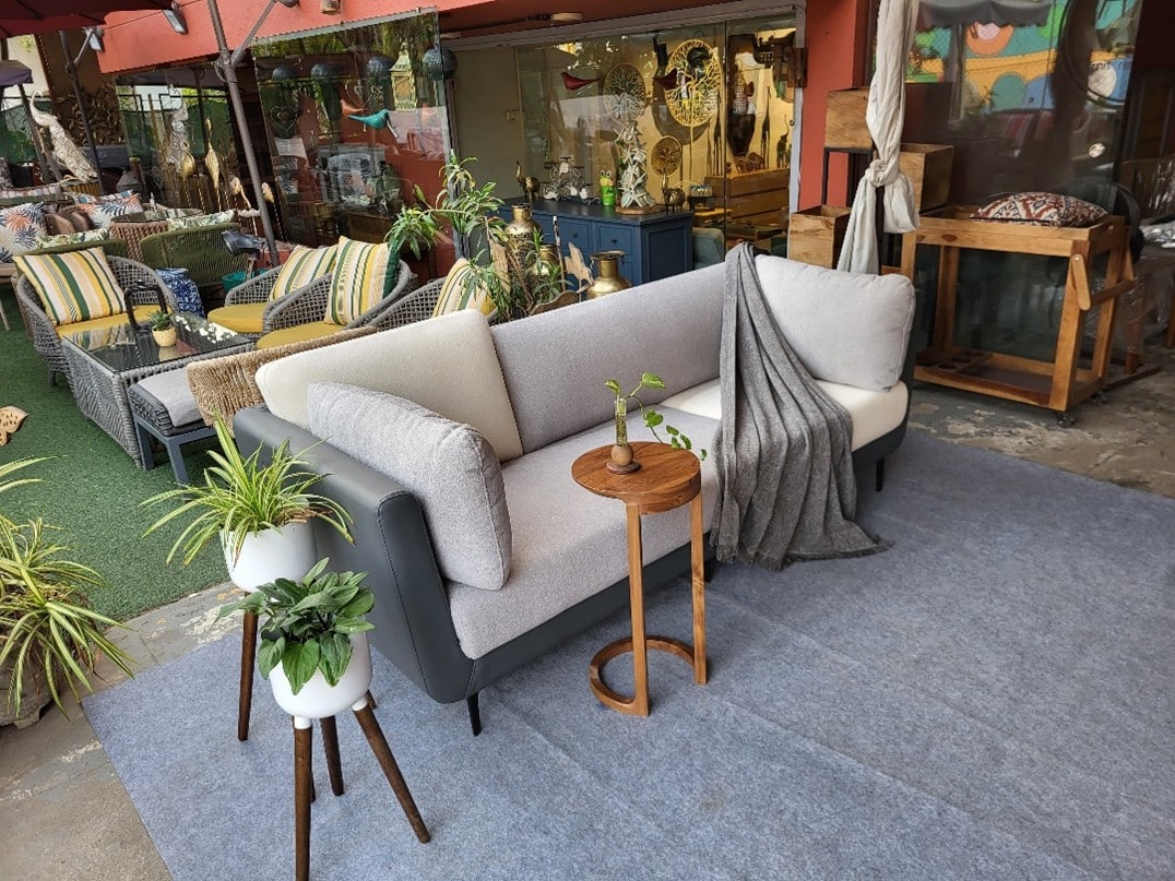 A stylish Teak wood sofa displayed in an outdoor showroom setting. The sofa features a modern, curved design with light grey fabric upholstery and a dark grey outer frame. A C-shaped teak wood side table holds a small plant, and a cozy grey throw is draped over the arm.